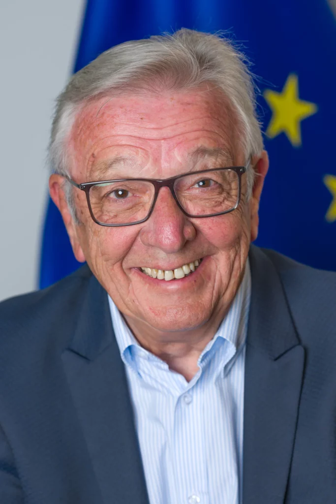 Professional portrait of a smiling man wearing glasses and a blue blazer, with a European Union flag behind him.