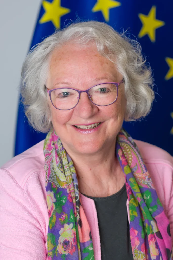 Professional portrait of a smiling woman wearing purple glasses, a pink jacket and a colourful scarf, with a European Union flag behind her.