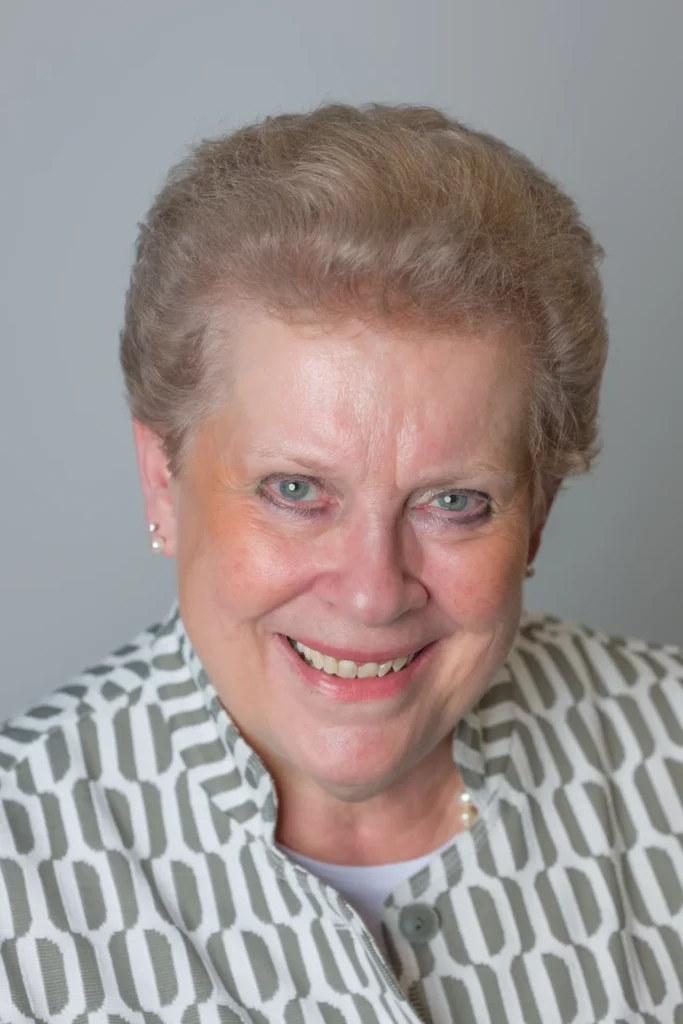 Professional portrait of Catherine Trautmann smiling, wearing pearl earrings and a patterned jacket against a grey background.