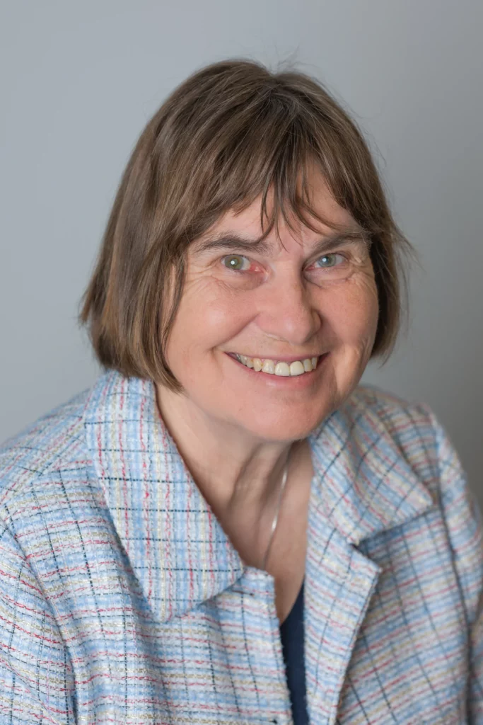 Professional portrait of a smiling woman with a brown bob haircut, wearing a light blue textured jacket against a grey background.