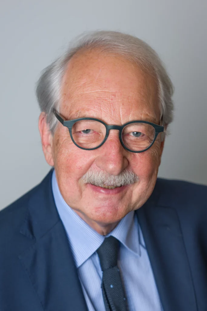 Portrait professionnel d'un homme portant des lunettes rondes et une moustache grise, vêtu d'un costume sombre et d'une cravate, sur fond gris.