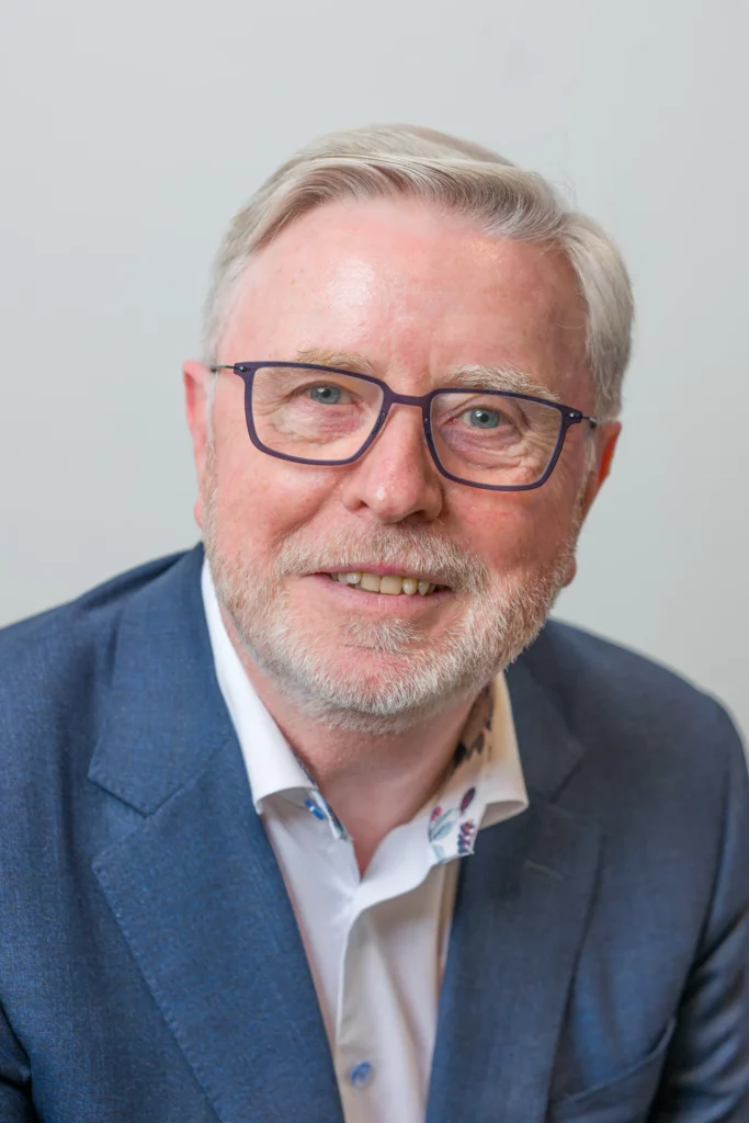 Professional portrait of a smiling man with glasses and a short grey beard, wearing a blue blazer and an open-collar white shirt.