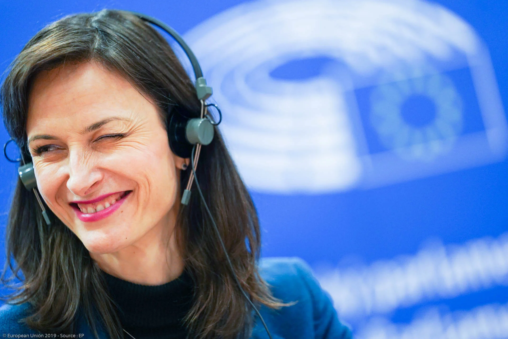Mariya Gabriel smiles whilst wearing an interpreting headset, in front of a sign at the European Parliament.