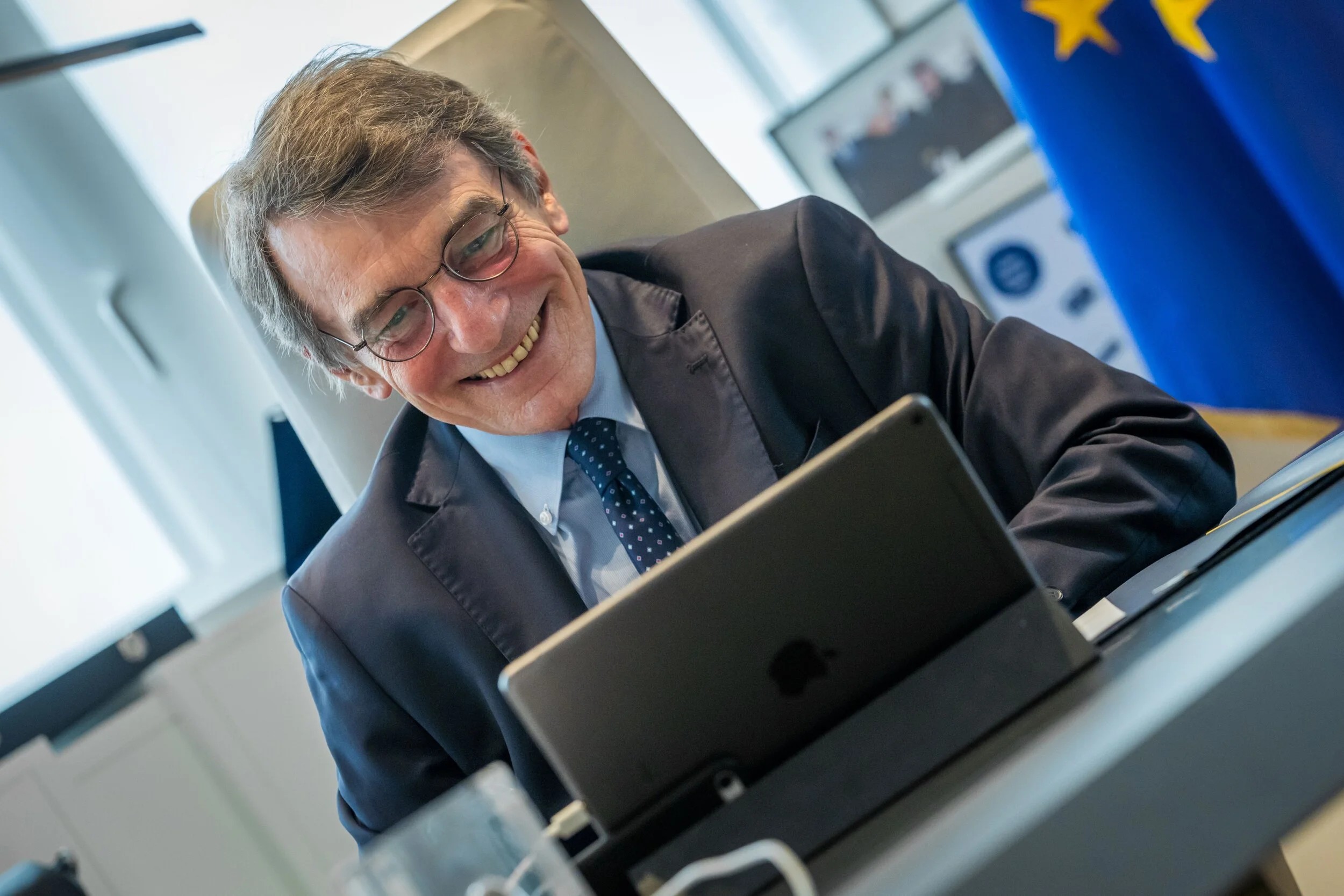 Homme souriant en costume, portant des lunettes, sur une tablette, drapeau de l'UE en arrière-plan.