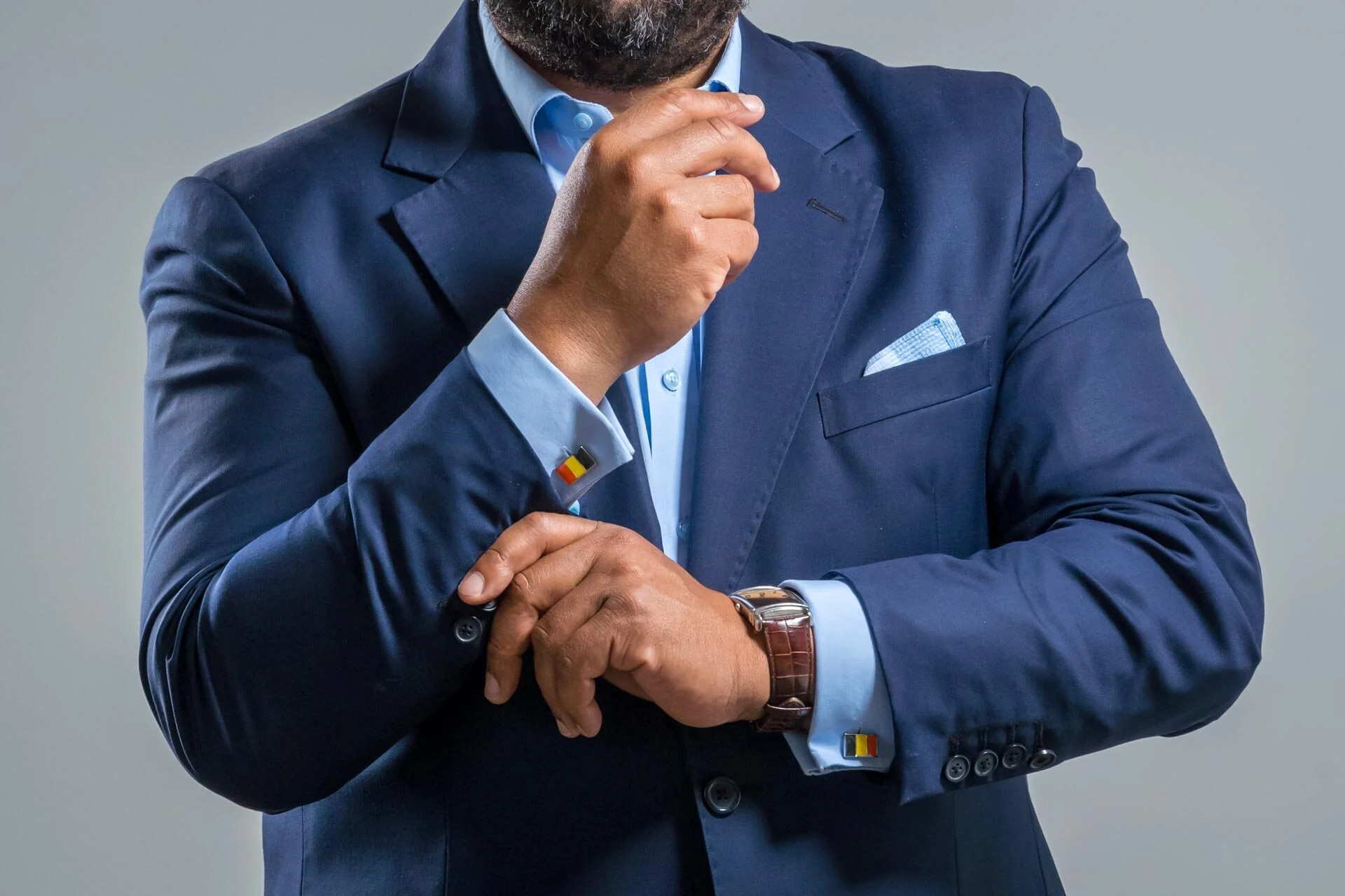Homme en costume bleu marine ajustant son bouton de manchette, portant une montre en cuir et une chemise bleue claire.