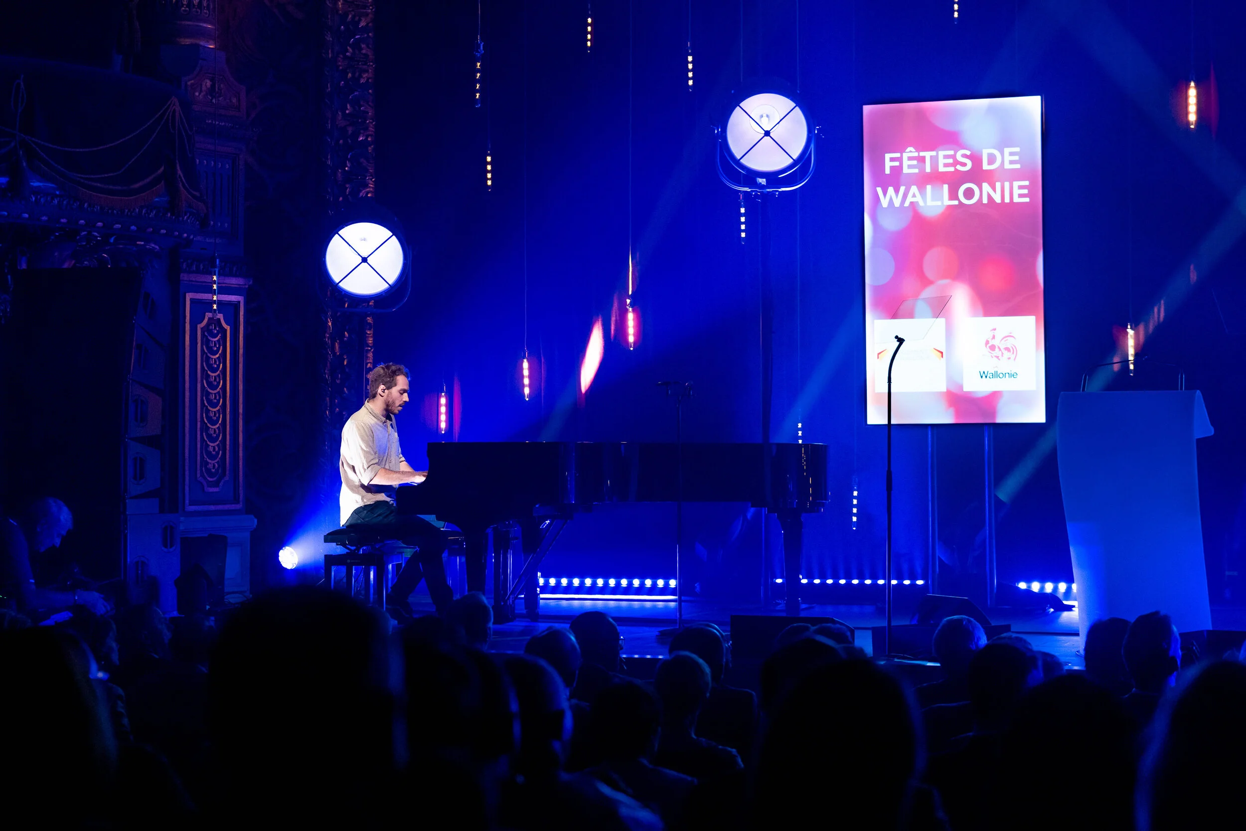 A pianist is playing on a stage lit up in blue, in front of the audience and a large screen.