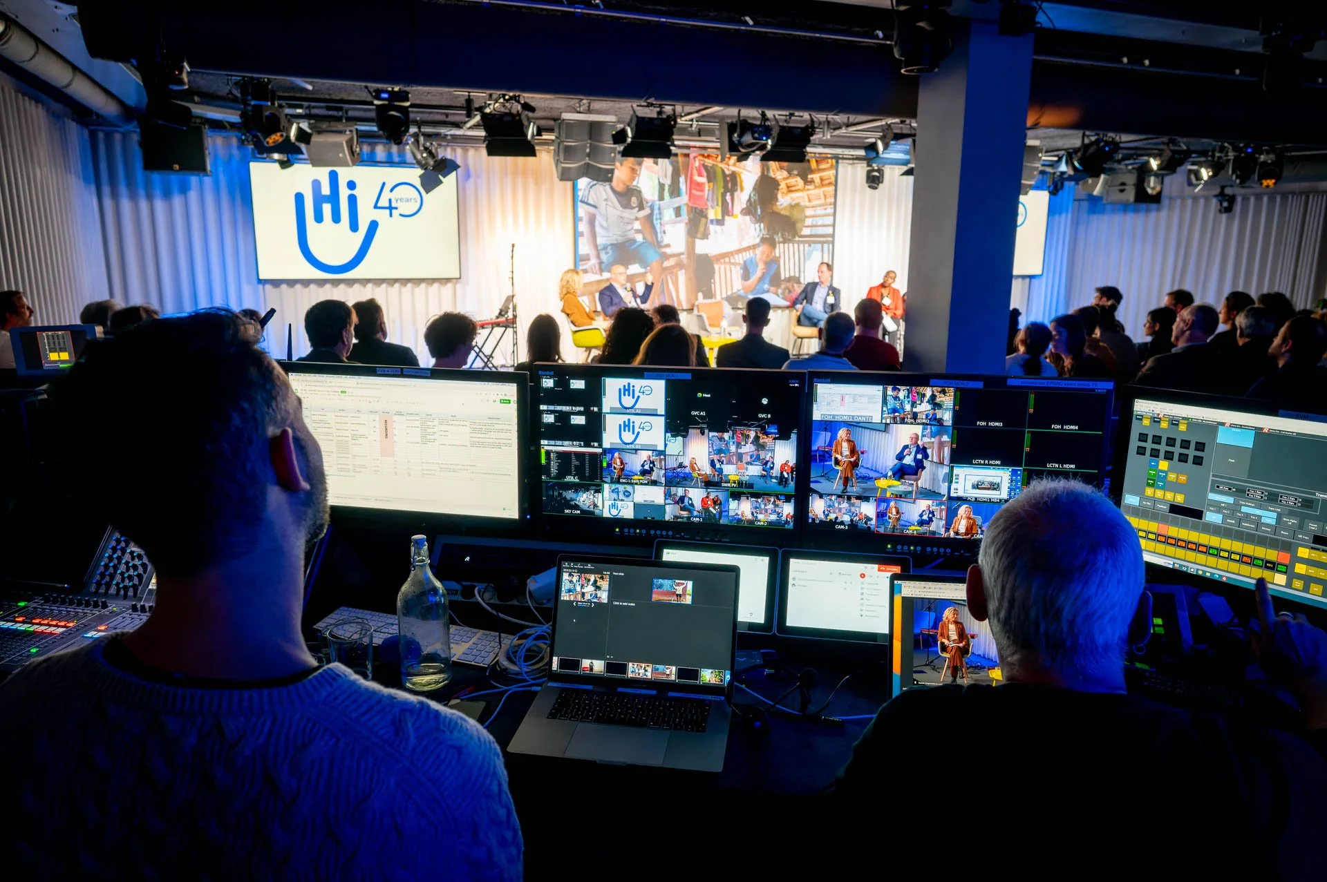 Vue depuis la régie audiovisuelle d’un événement, avec des écrans de contrôle au premier plan et une scène éclairée à l’arrière-plan.
