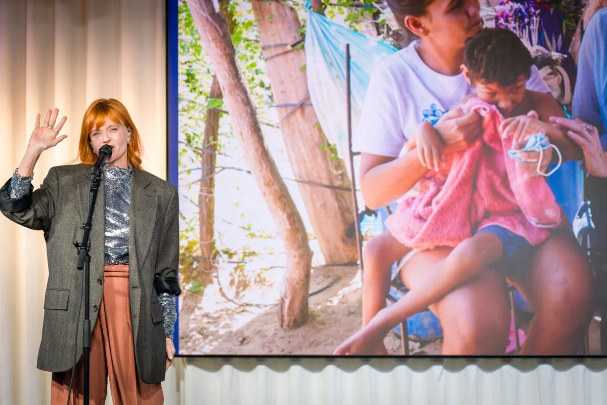 A woman with red hair, wearing a grey blazer and salmon-coloured trousers, is speaking into a microphone during a presentation, with a large poster in the background showing children and adults in a lively