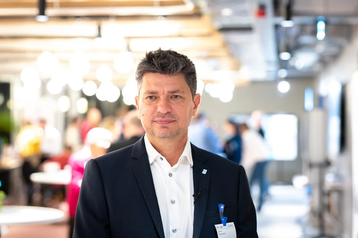 A man in a black suit and white shirt, wearing a name badge, is standing in an office or meeting room with people blurred in the background.