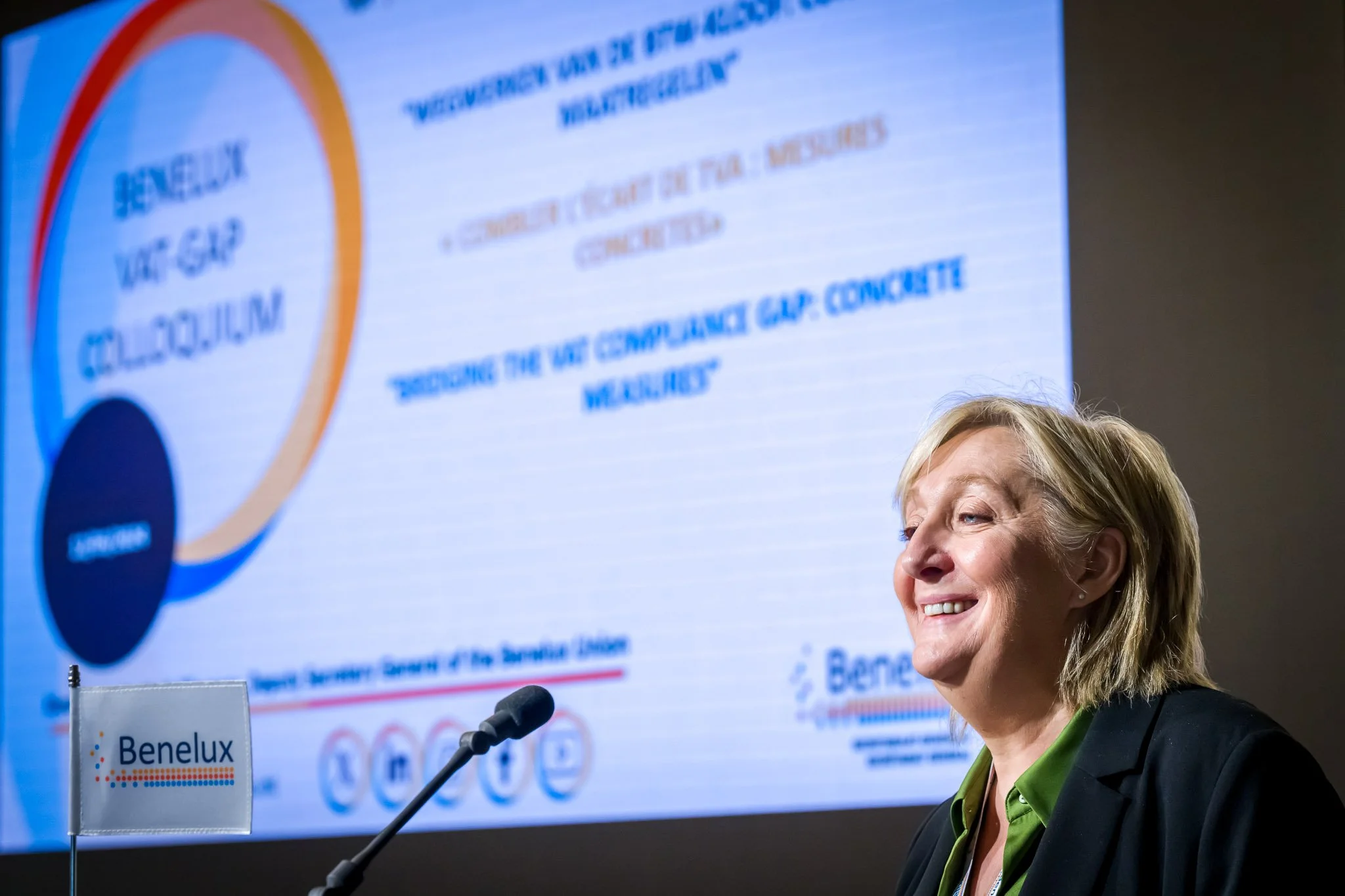 Une femme souriante lors d'une présentation au colloque Benelux sur le "VAT Gap" avec un écran en arrière-plan affichant des informations sur l'événement.