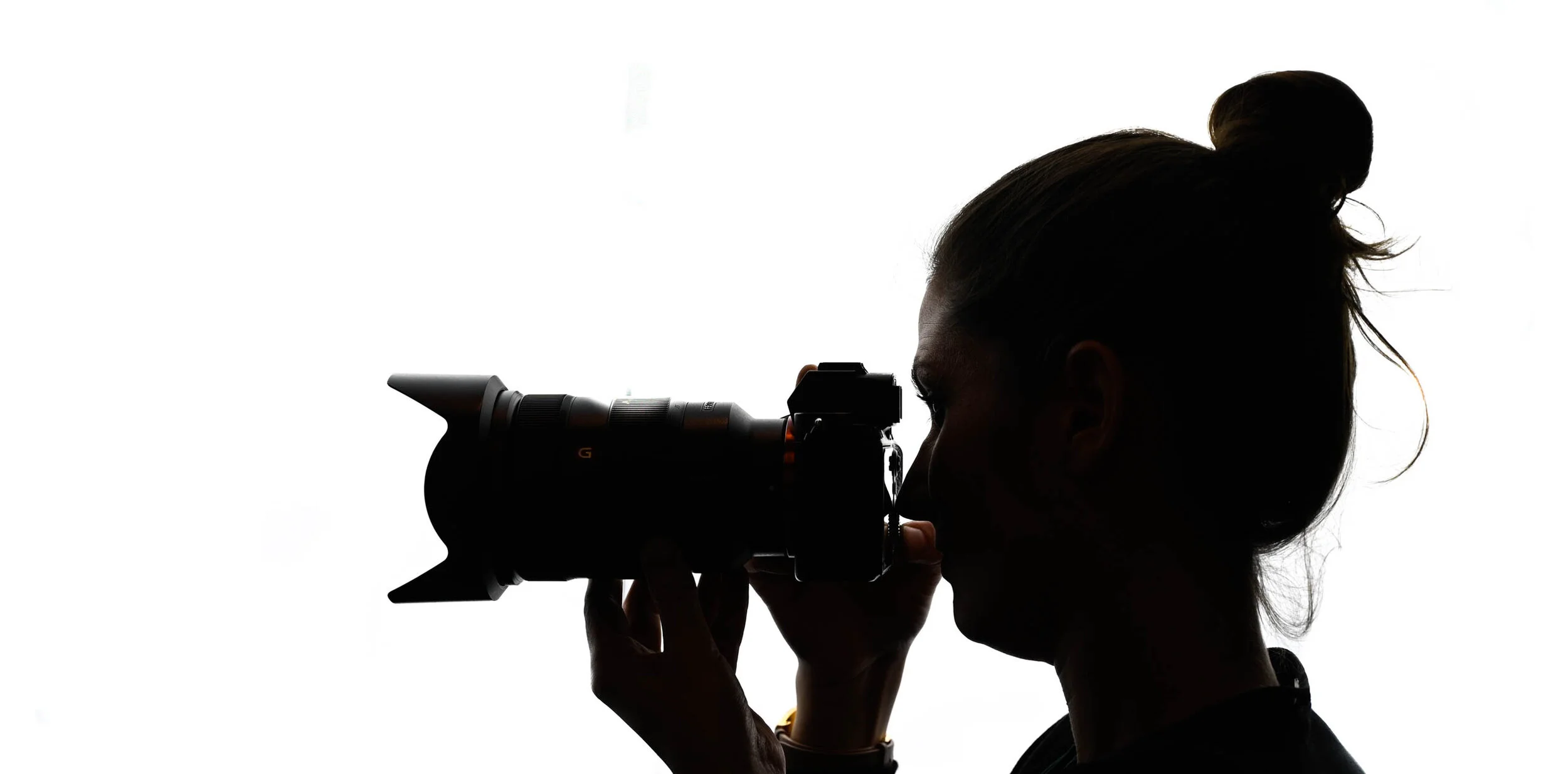 A silhouette of a female photographer with her hair tied back, looking through the viewfinder of her camera against a white background.