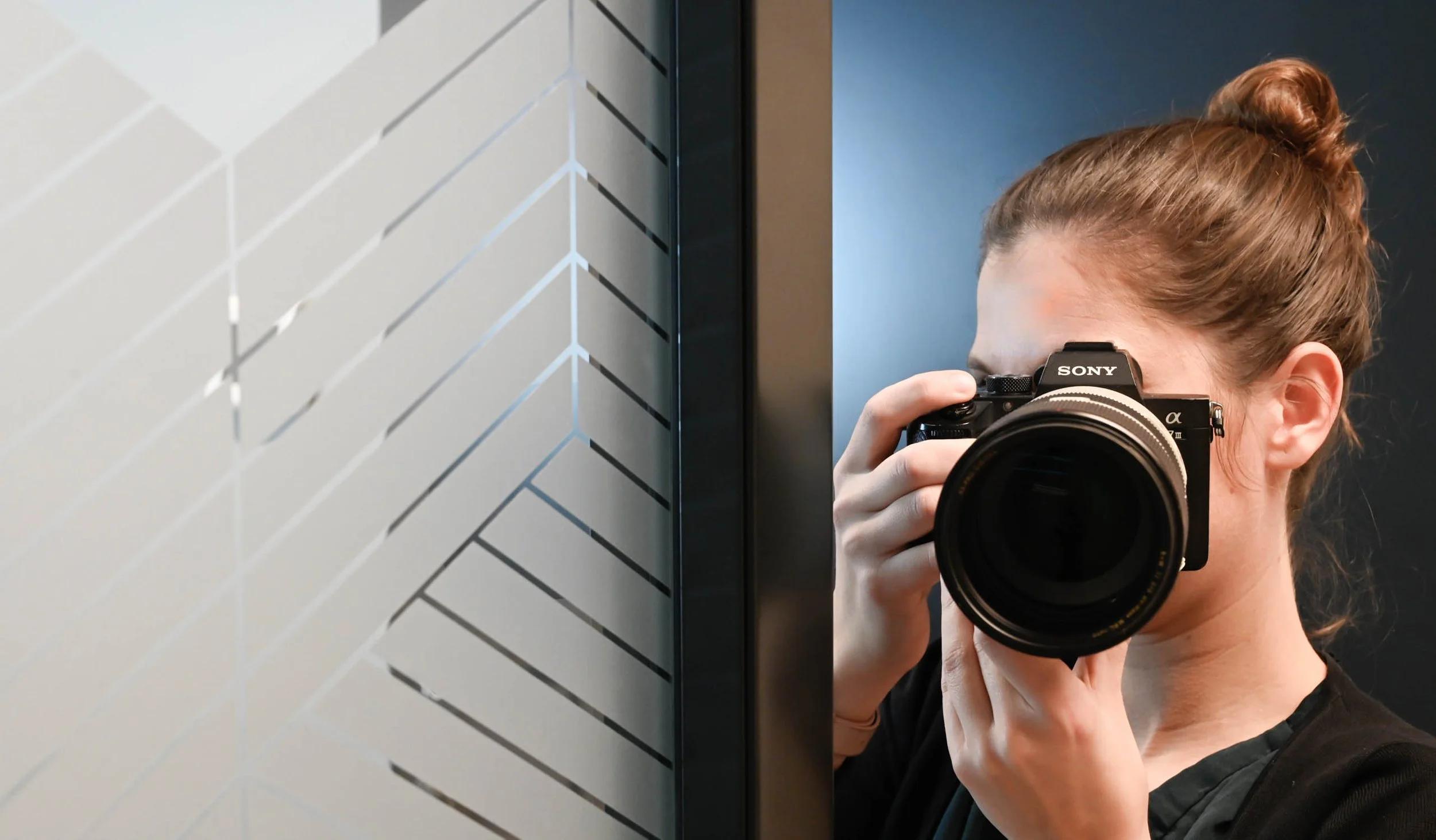 A photographer with her hair tied back is holding her camera in front of her face, near a wall with geometric patterns.