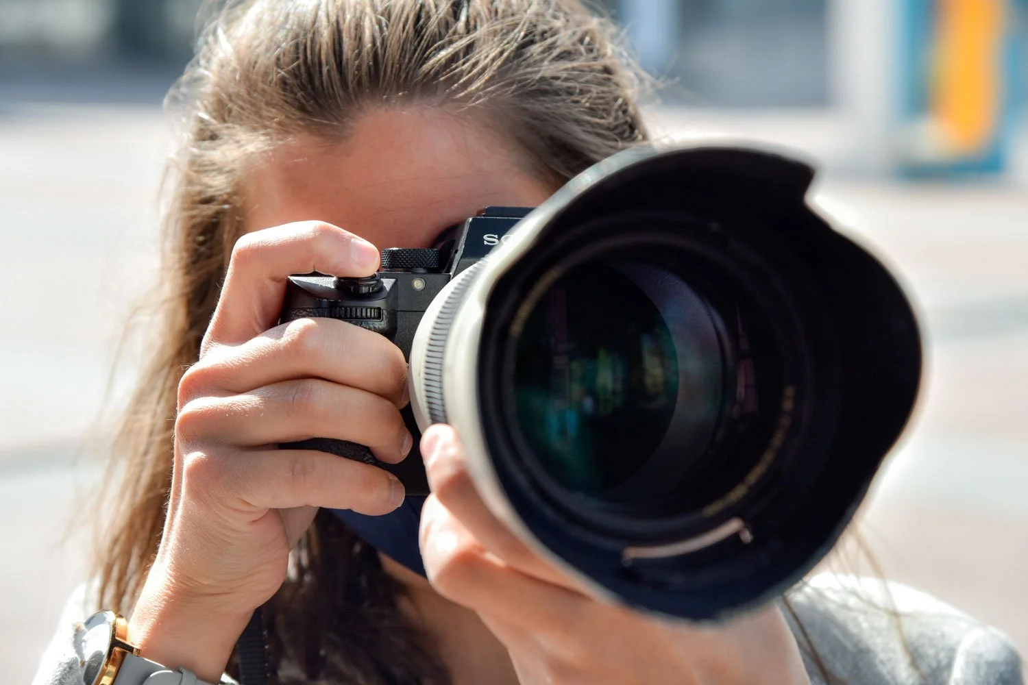 A close-up of a photographer looking through the viewfinder of her camera, with the lens pointing towards the viewer.