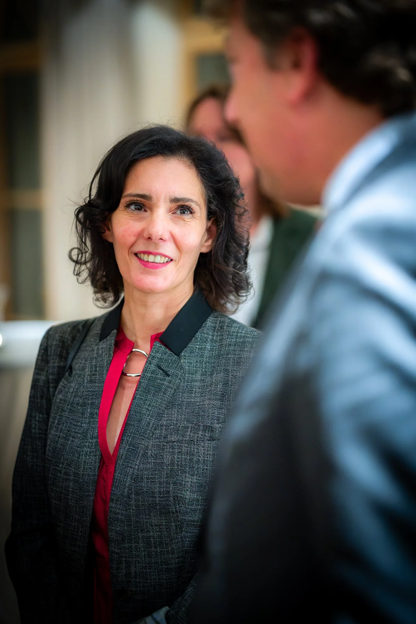 A woman with dark hair and a grey jacket smiles while speaking with a man at an indoor event.