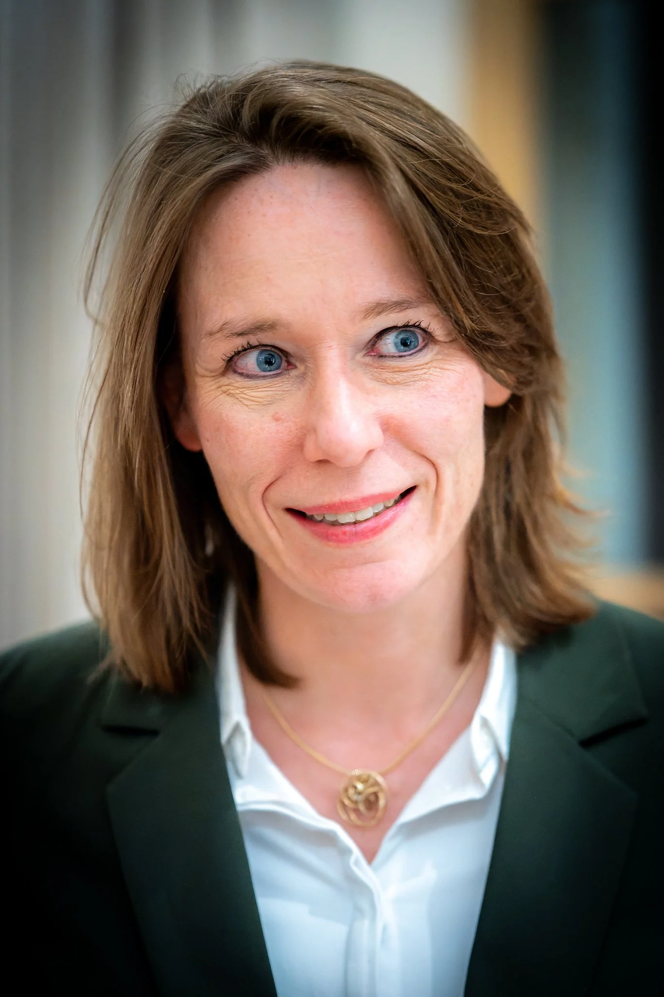 A smiling woman with brown hair, wearing a green jacket and a gold necklace.