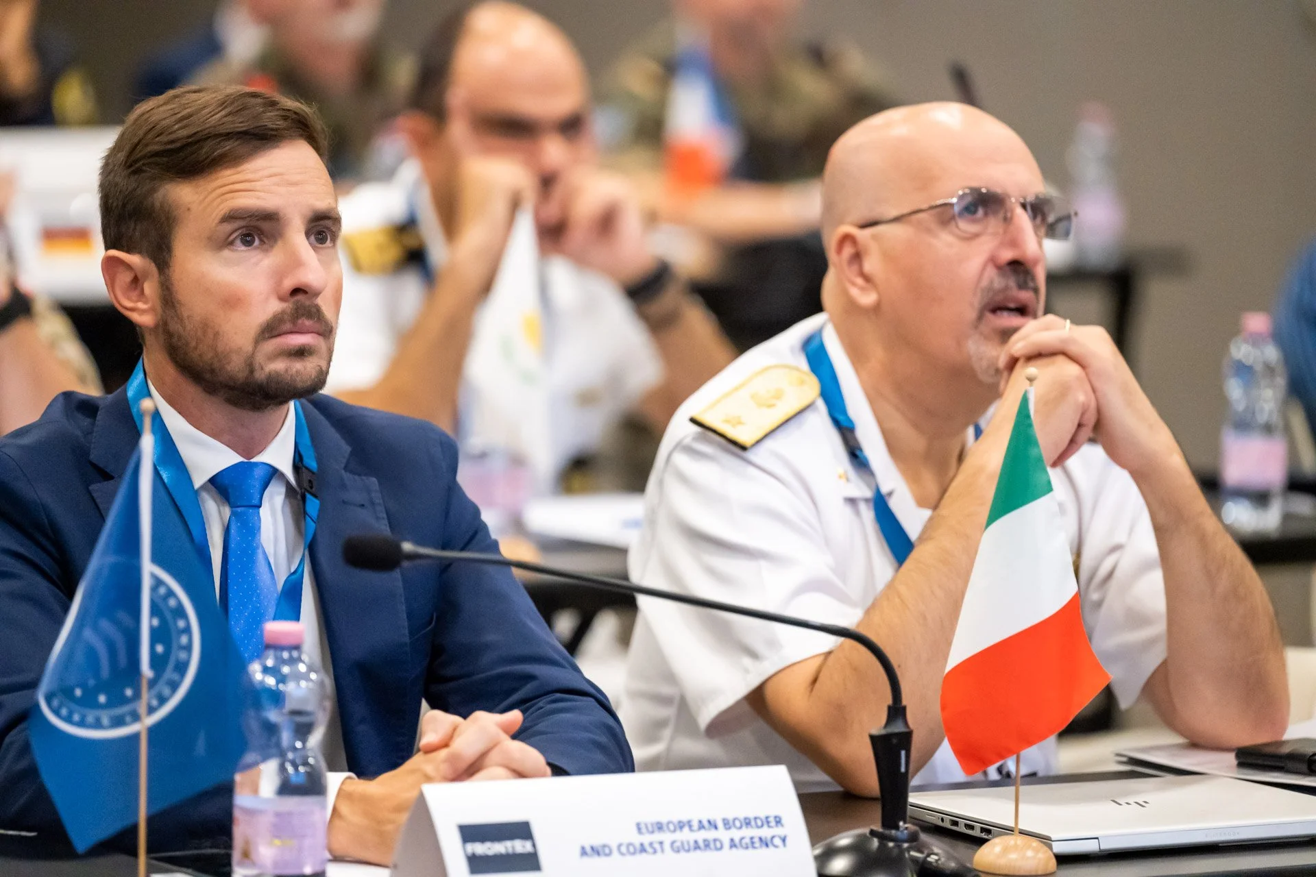 Deux hommes assis à une conférence, avec des drapeaux et micros devant eux.