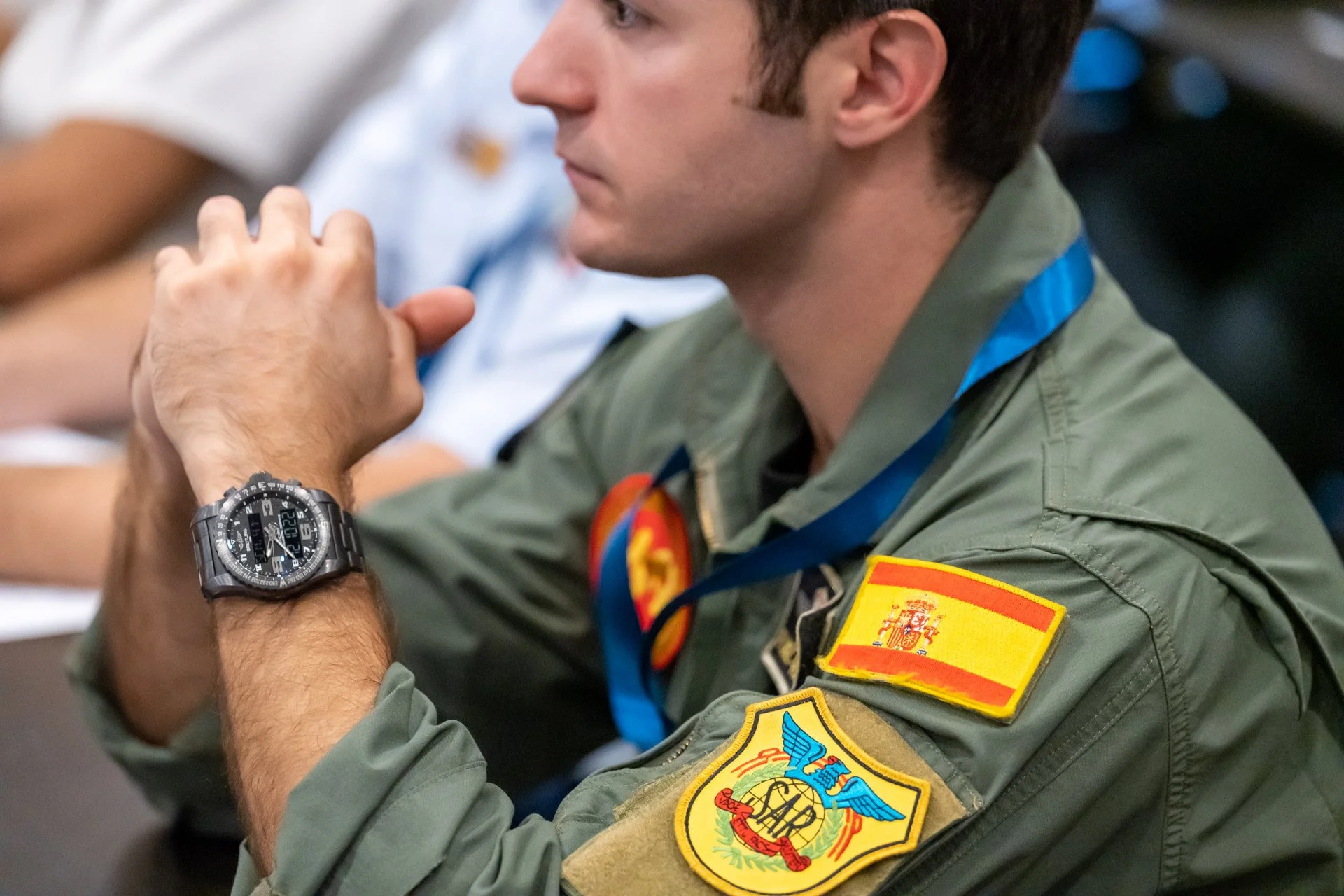 Un homme en uniforme militaire avec un écusson espagnol sur la manche, assis à une table. Il porte une montre-bracelet et un insigne sur la veste. Le fond est flou.