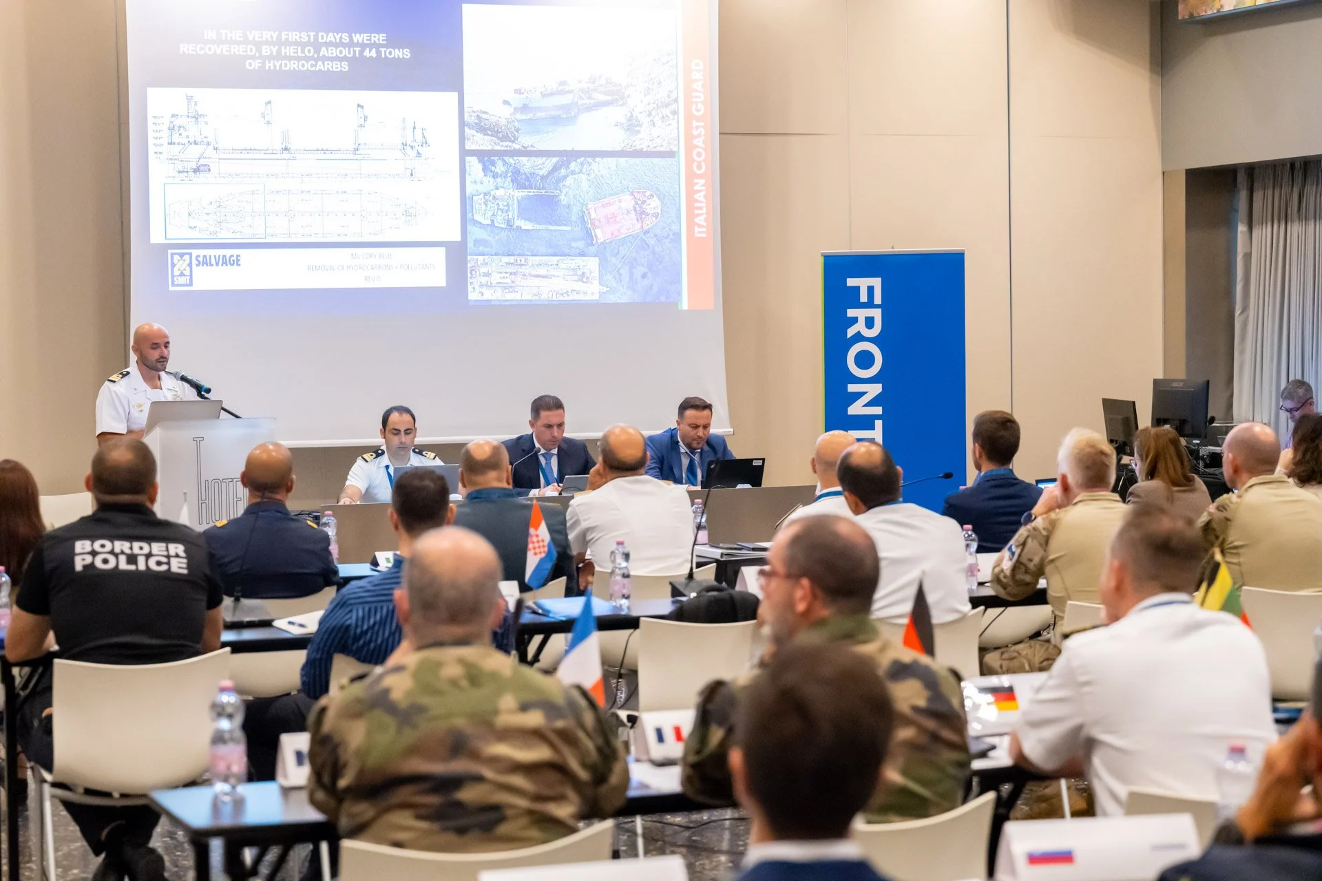 Photo d'une conférence avec des officiers militaires et des représentants de la garde côtière, un écran projetant des informations."
