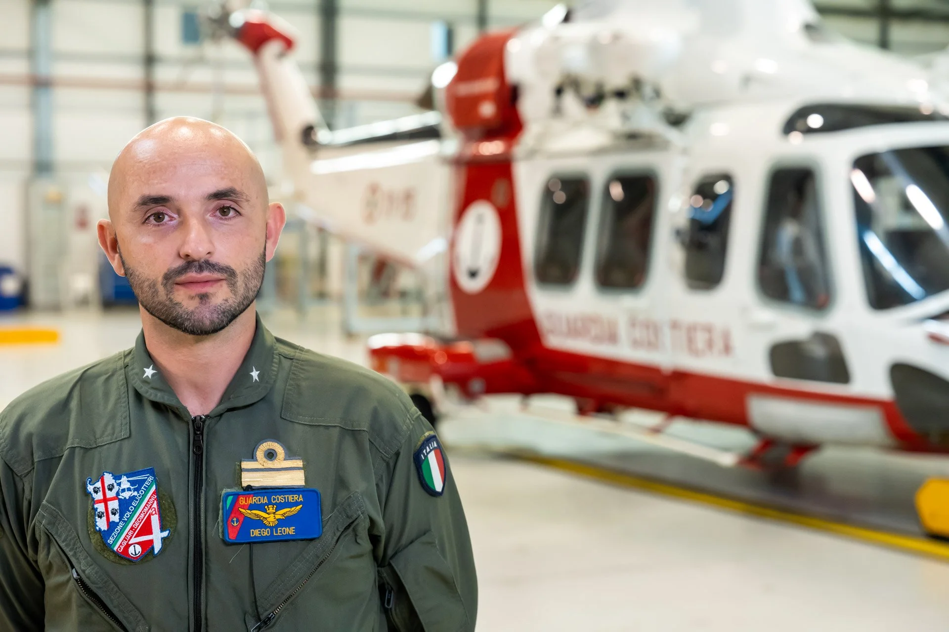 Un homme en uniforme militaire avec un insigne "Guardia Costiera" se tient devant un hélicoptère rouge et blanc à l'intérieur d'un hangar.