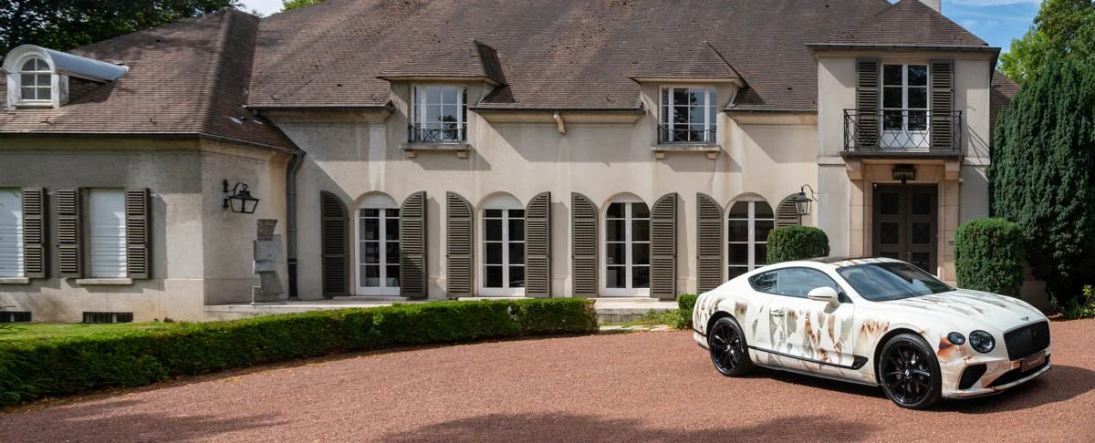 Voiture de luxe blanche devant une grande maison élégante avec des fenêtres à volets, allée en gravier, et jardin bien entretenu.