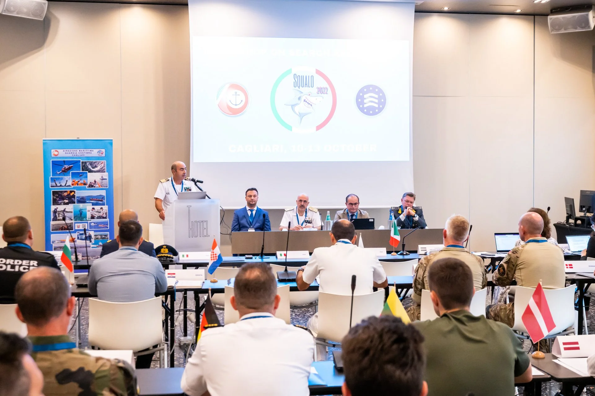 Réunion militaire internationale avec des représentants en uniforme dans une salle de conférence, drapeaux nationaux sur les tables, présentation sur un écran, "Squalo 2022" écrit avec les dates 10-13 octobre à Cagliari.