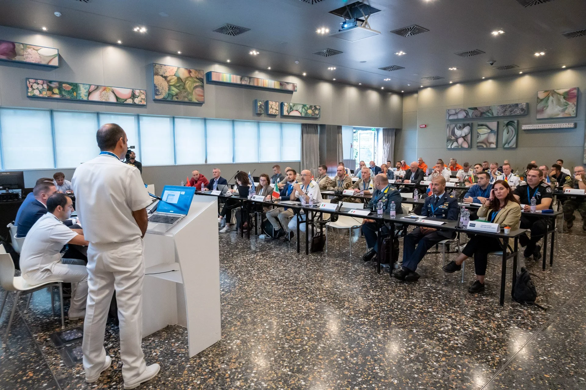 Un intervenant debout présente un exposé à des participants assis à des tables lors d’une réunion liée à une opération Frontex.