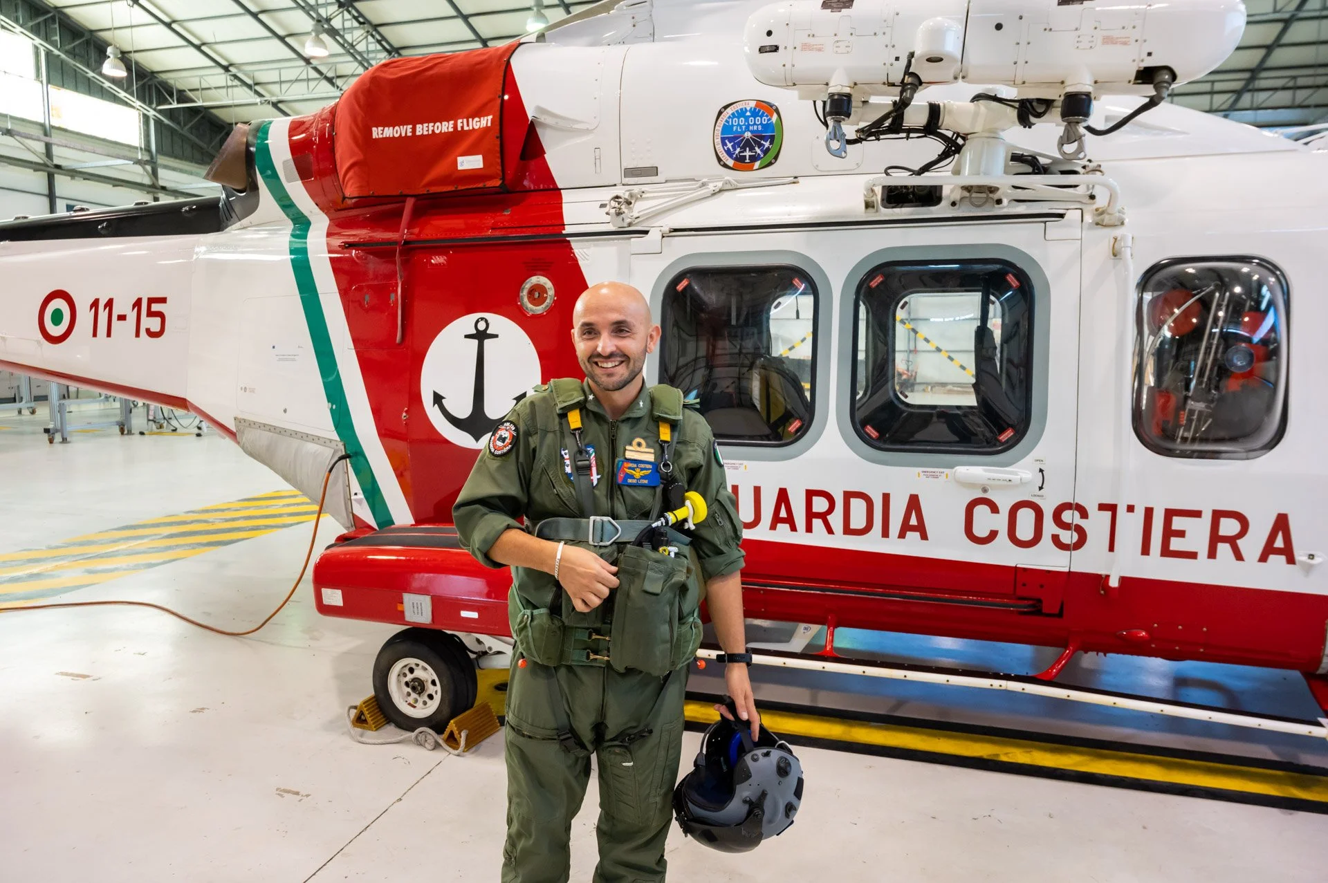 Un pilote souriant se tient devant un hélicoptère de la Garde côtière, portant une combinaison de vol et tenant un casque dans un hangar.