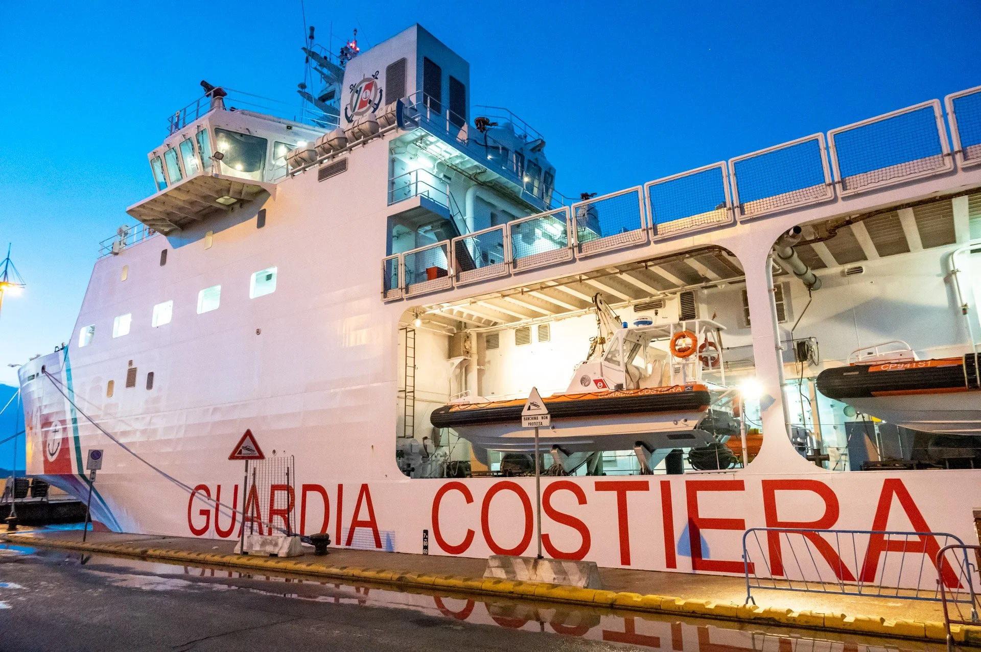 Navire de la garde côtière italienne en quai, avec des canots de sauvetage à bord, en soirée.