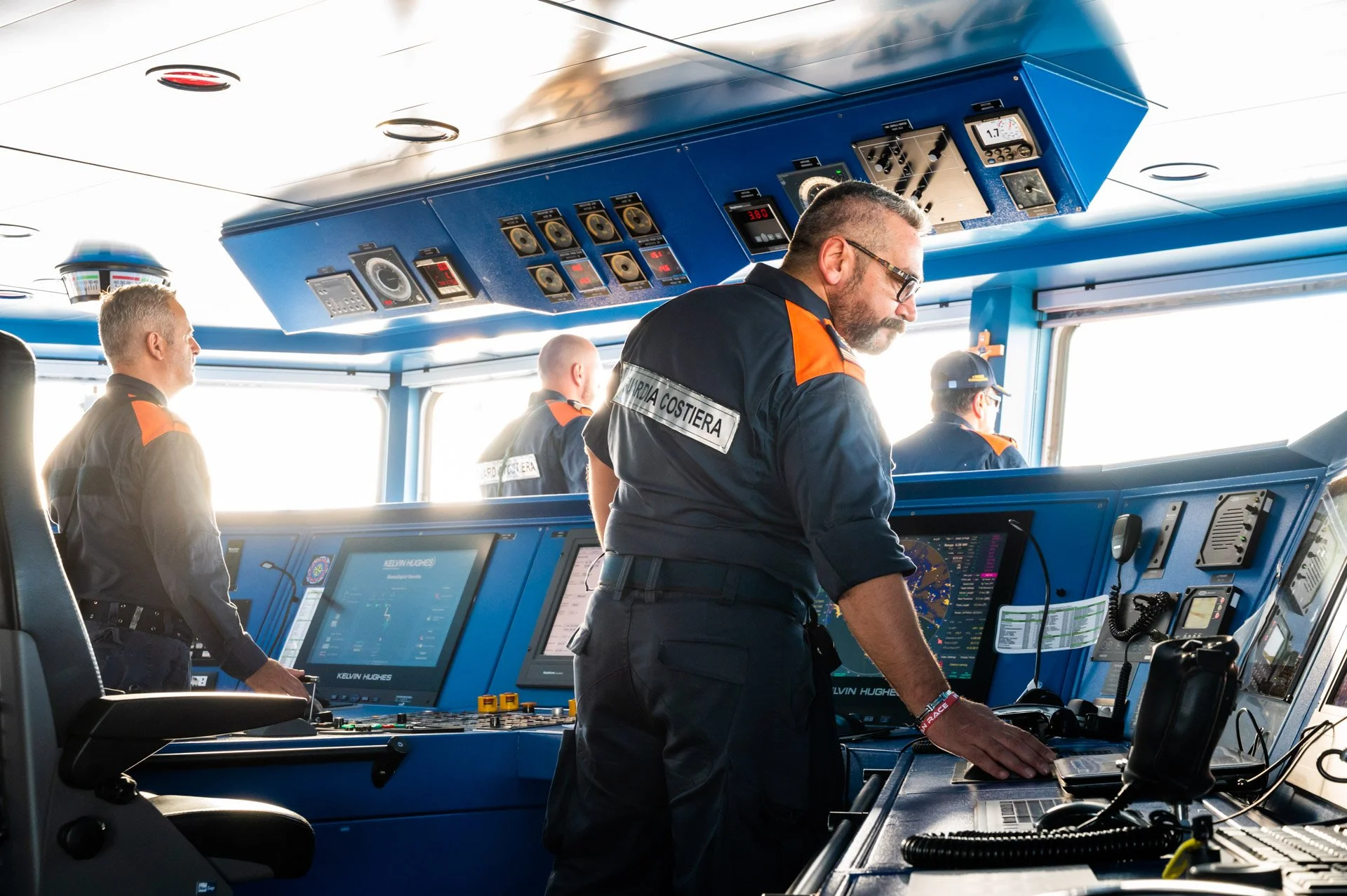 Quatre personnes en uniforme se trouvent dans une salle de contrôle de navire, manipulant des équipements de navigation.