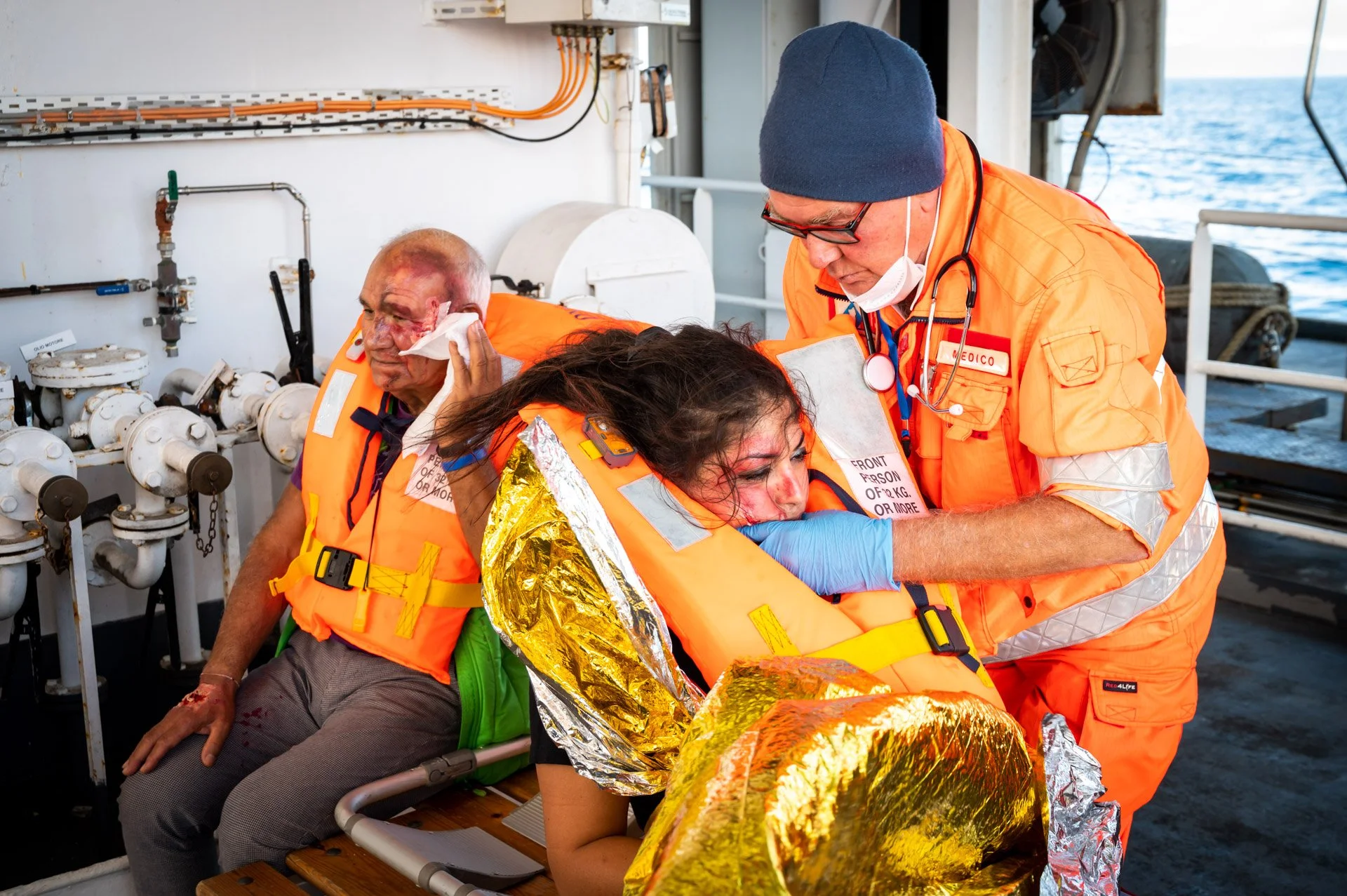 Scène de secours en mer, avec un sauveteur en tenue orange aidant deux personnes portant des gilets de sauvetage, dont une enveloppée dans une couverture de survie.