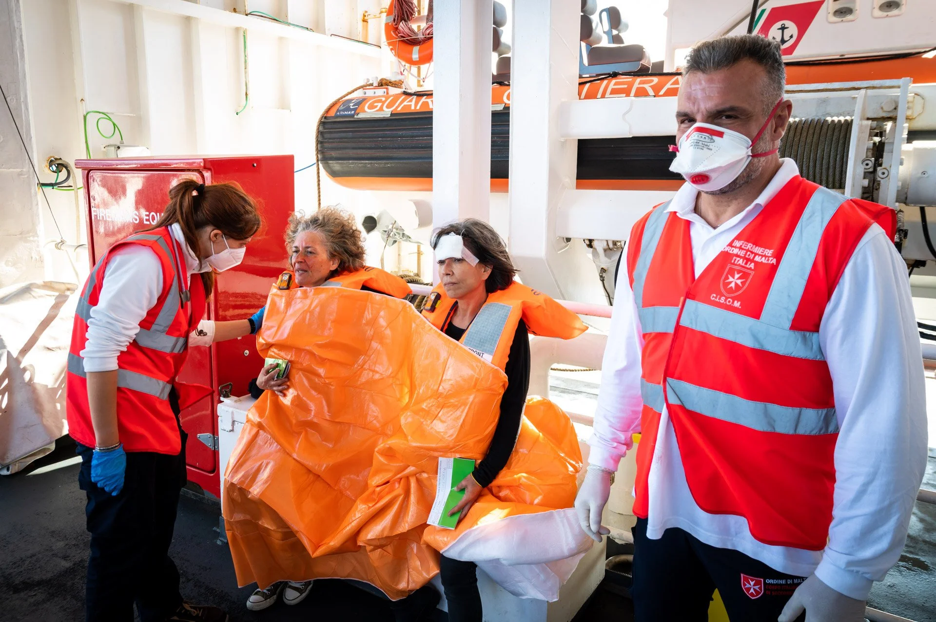 Quatre personnes sur un bateau, dont deux femmes assises portant des couvertures de survie orange et des gilets de sauvetage, et deux membres du personnel de secours portant des gilets de sécurité rouges et des masques.