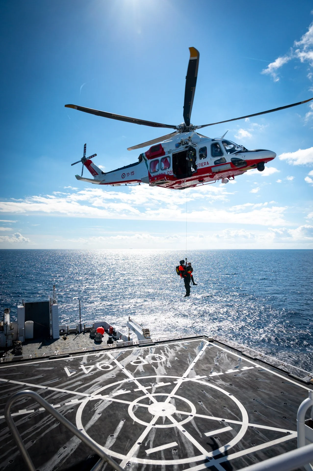 Photographie corporate d'un hélicoptère de sauvetage en mer avec une personne descendant en tyrolienne, au-dessus de l'eau, près d'un navire avec hélipad.
