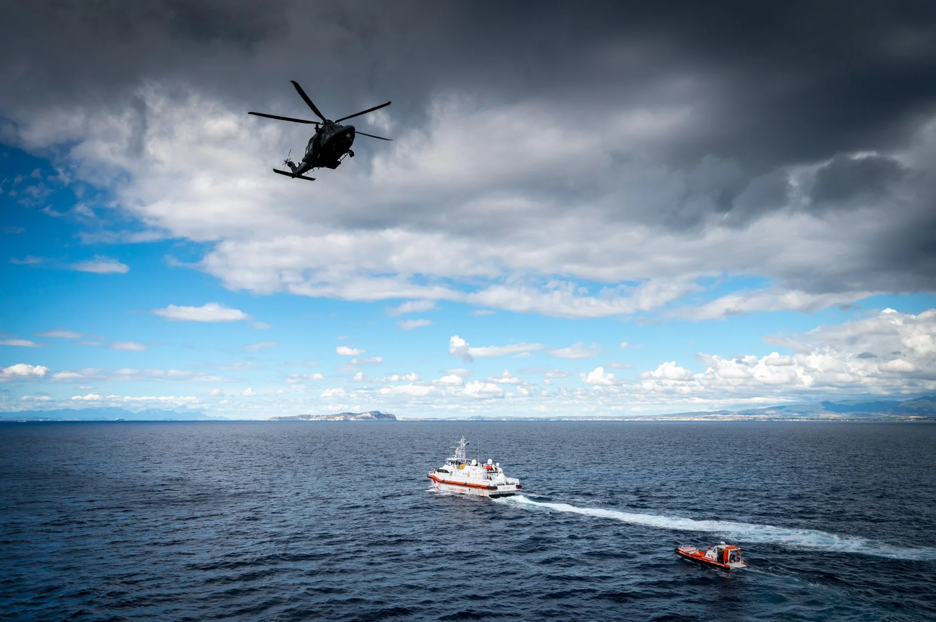 Hélicoptère planant au-dessus de la mer, bateau de sauvetage orange et blanc sur l'eau, ciel nuageux.