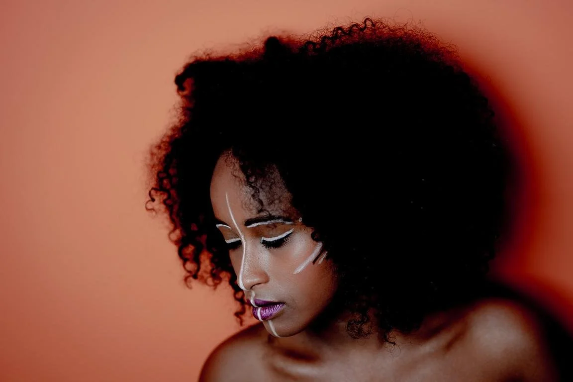 Femme avec cheveux bouclés et maquillage artistique blanc sur fond orange.