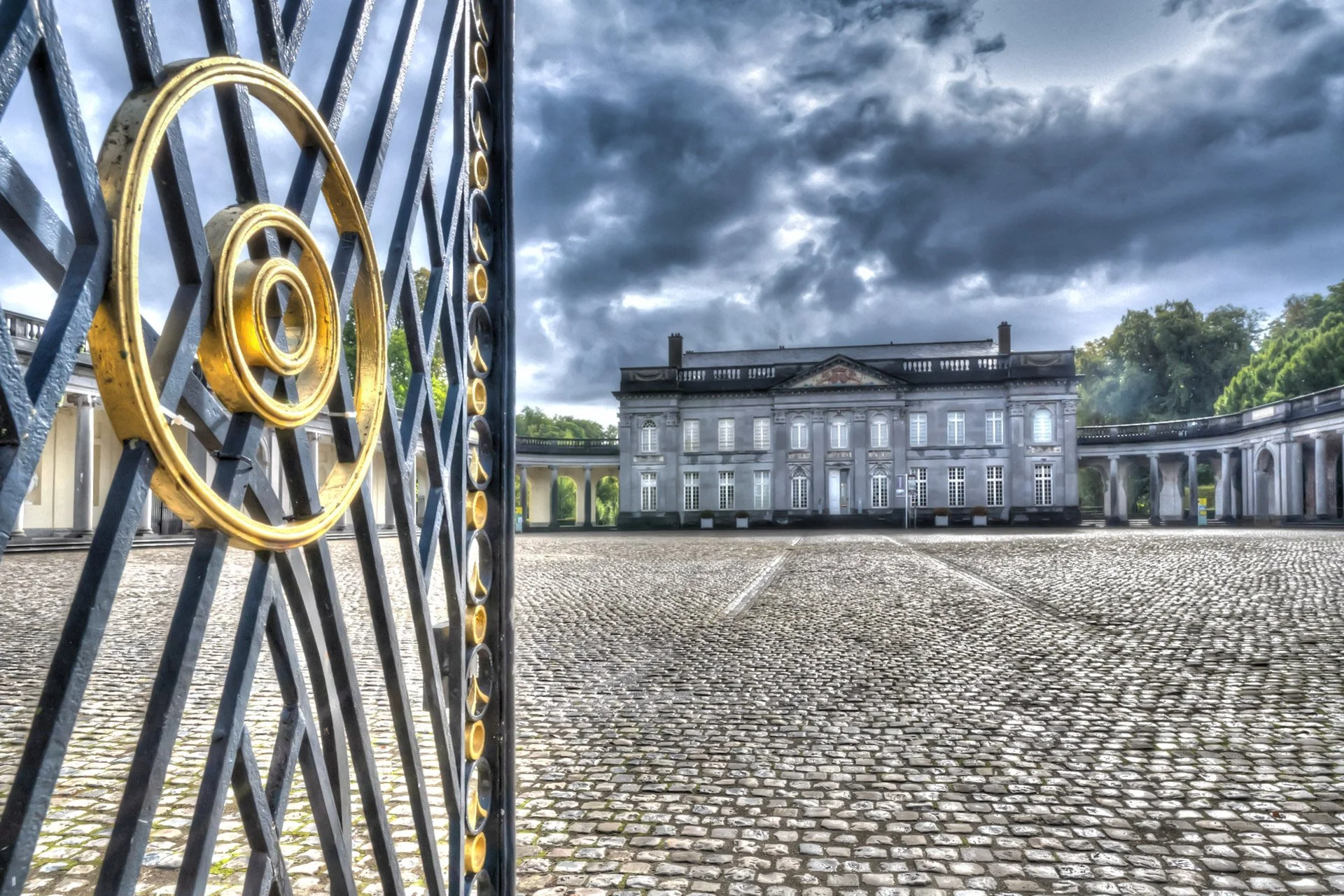 Le château de Seneffe vu depuis sa cour pavée, avec un portail en fer forgé orné de dorures au premier plan.