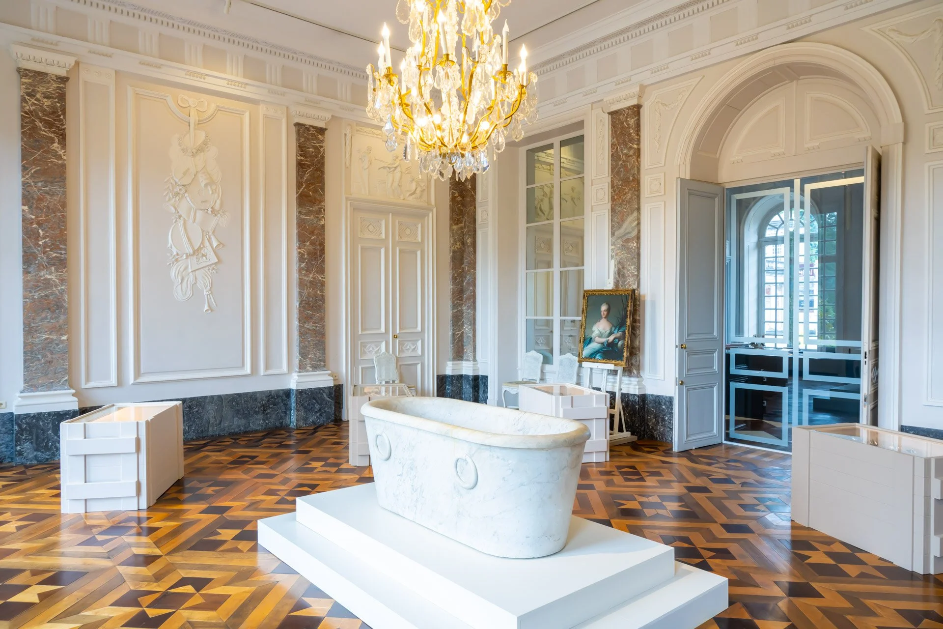Une salle du château de Seneffe avec un lustre doré, des moulures et un parquet à motifs, présentant des pièces d’orfèvrerie en vitrine.