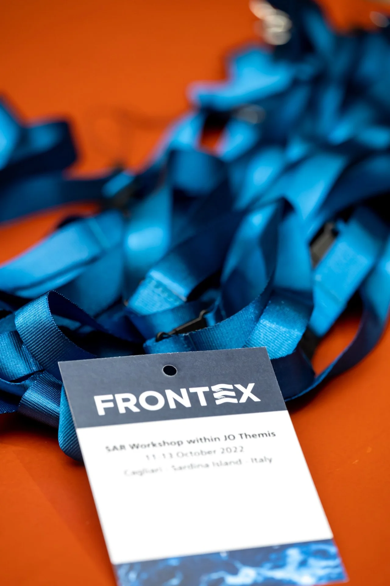 Badges de conférence Frontex avec cordons bleus sur une table orange, texte indiquant "SAR Workshop within JO Themis, 11-13 October 2022, Cagliari, Sardinia Island, Italy".