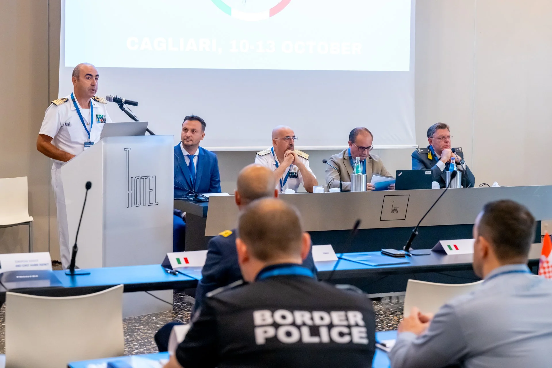Conférence avec des officiers en uniforme à une table, un homme parlant au podium, et une projection indiquant "Cagliari, 10-13 October."