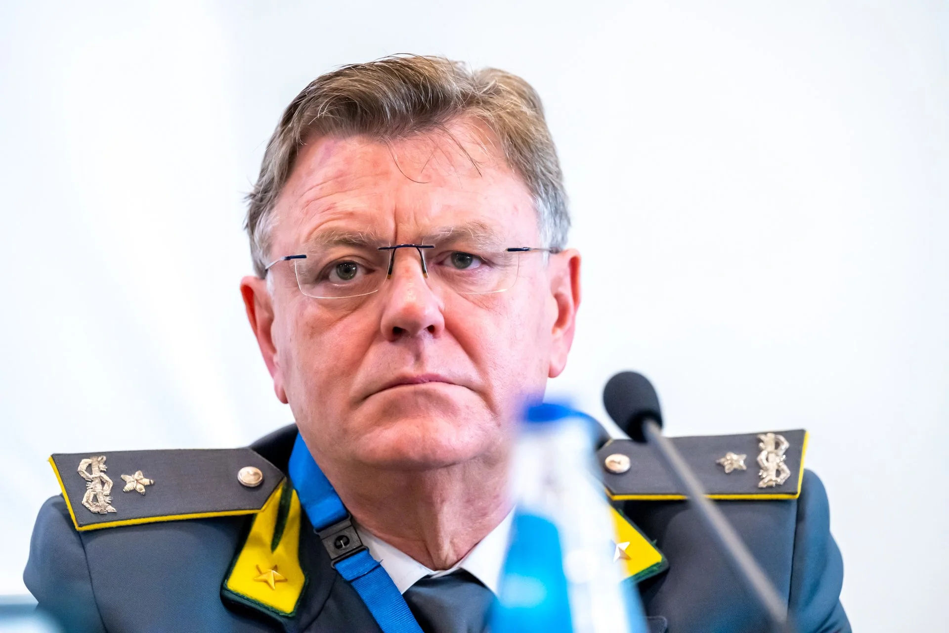 Un homme en uniforme avec des épaulettes, portant des lunettes, assis devant un micro.