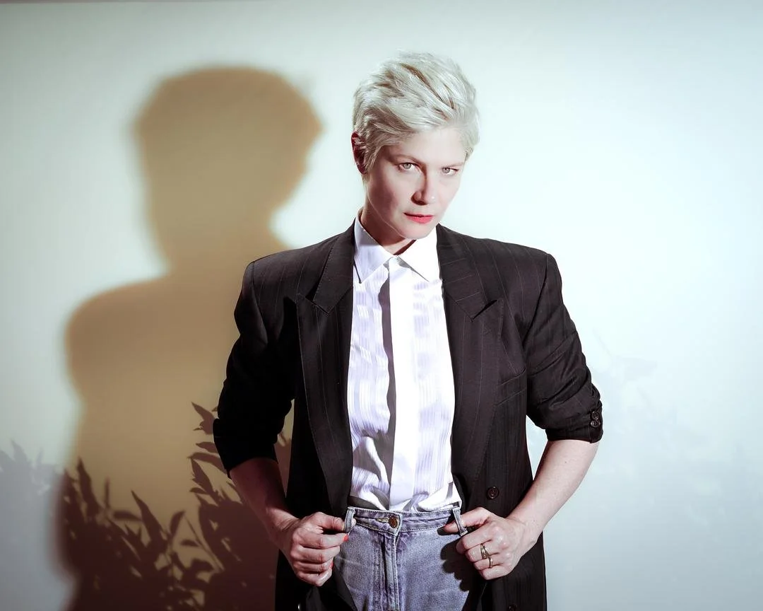 Une personne aux cheveux blonds courts porte un costume sombre avec une chemise blanche et une cravate. Elle est debout contre un mur clair avec son ombre projetée à l'arrière-plan.
