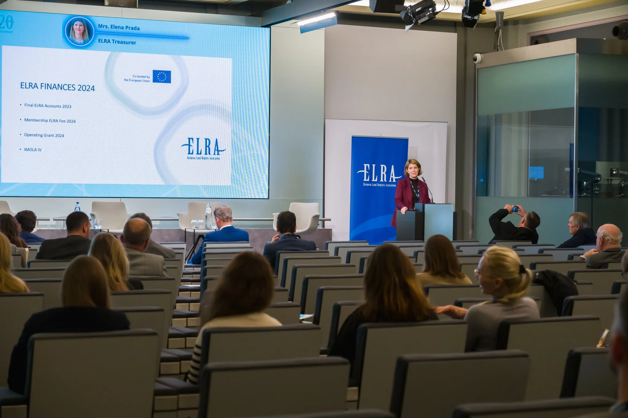 Photographie professionnelle d'une conférence sur les finances de l'ELRA 2024 avec une présentatrice debout près d'un pupitre, un écran affichant des informations financières, et un public assis dans une salle de conférence.