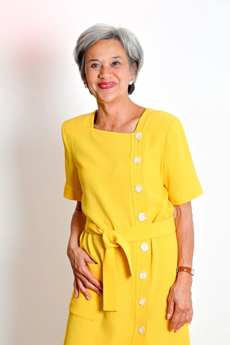 Une femme aux cheveux gris portant une robe jaune avec des boutons, souriant devant un fond blanc.