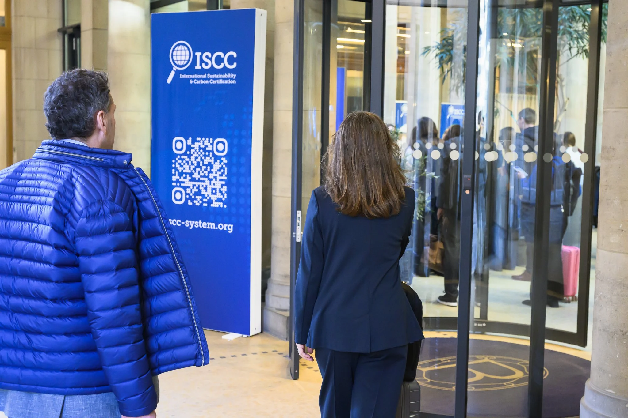 Two visitors approach a glass entrance beside a blue ISCC banner with a QR code.