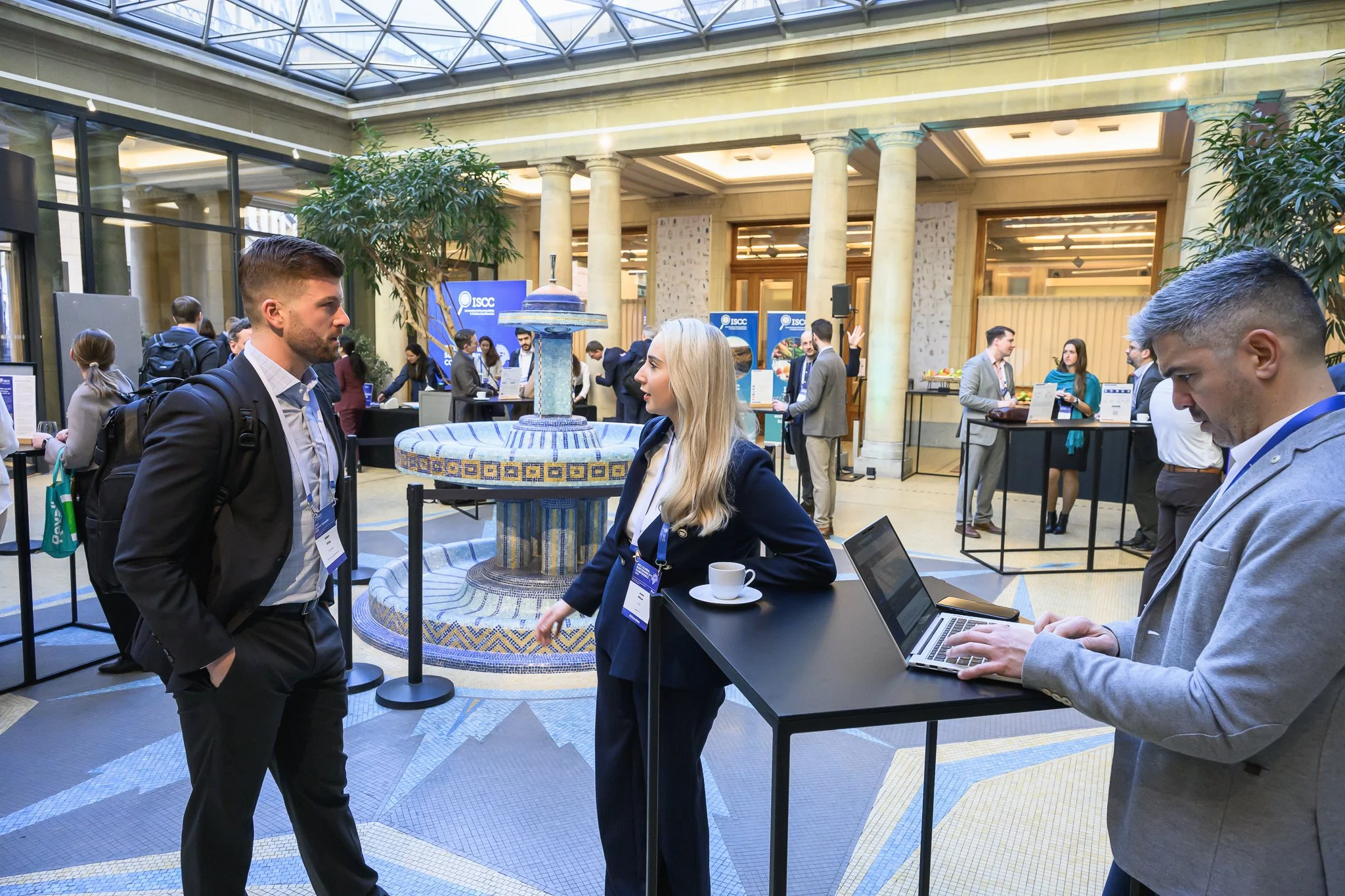 Participants stand around tall tables and look at their conference materials in a glass-roofed lobby.