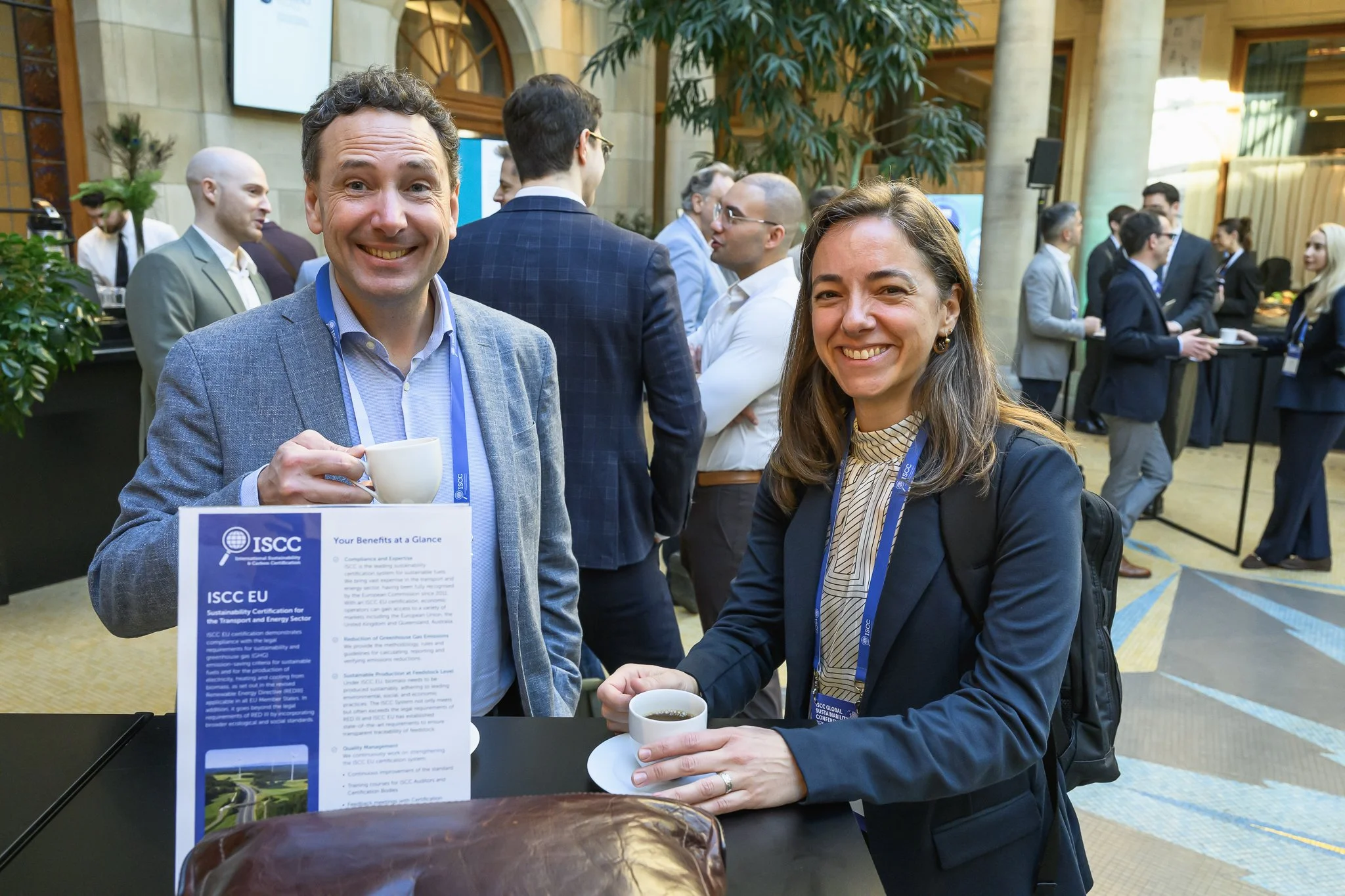 A smiling man and woman hold cups beside a conference information stand in the lobby.
