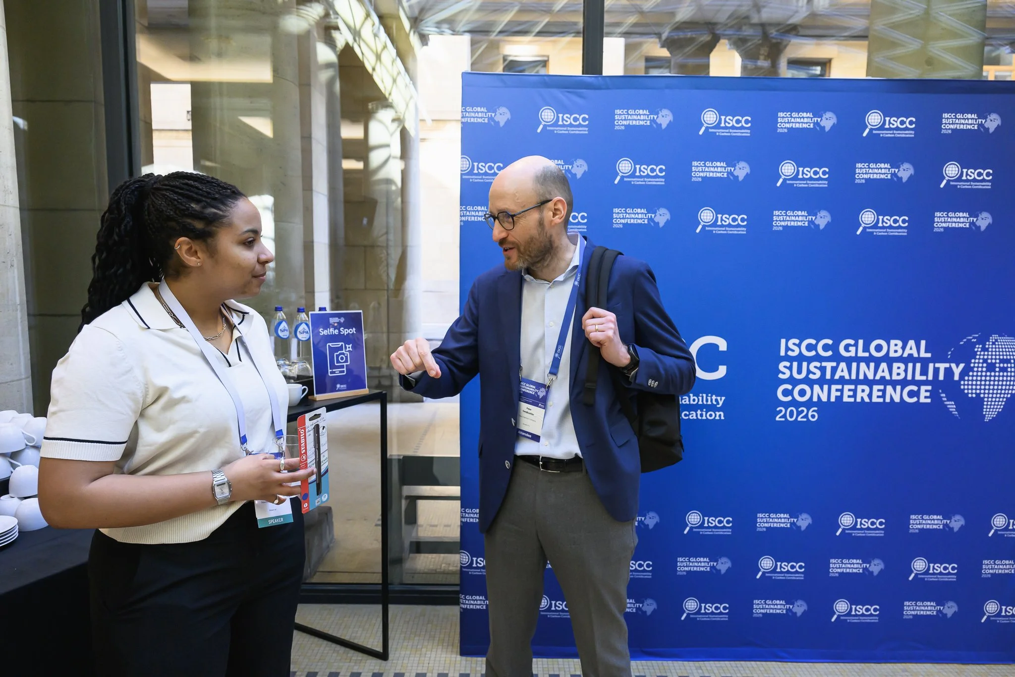 A woman and a man talk in front of an ISCC Global Sustainability Conference backdrop.