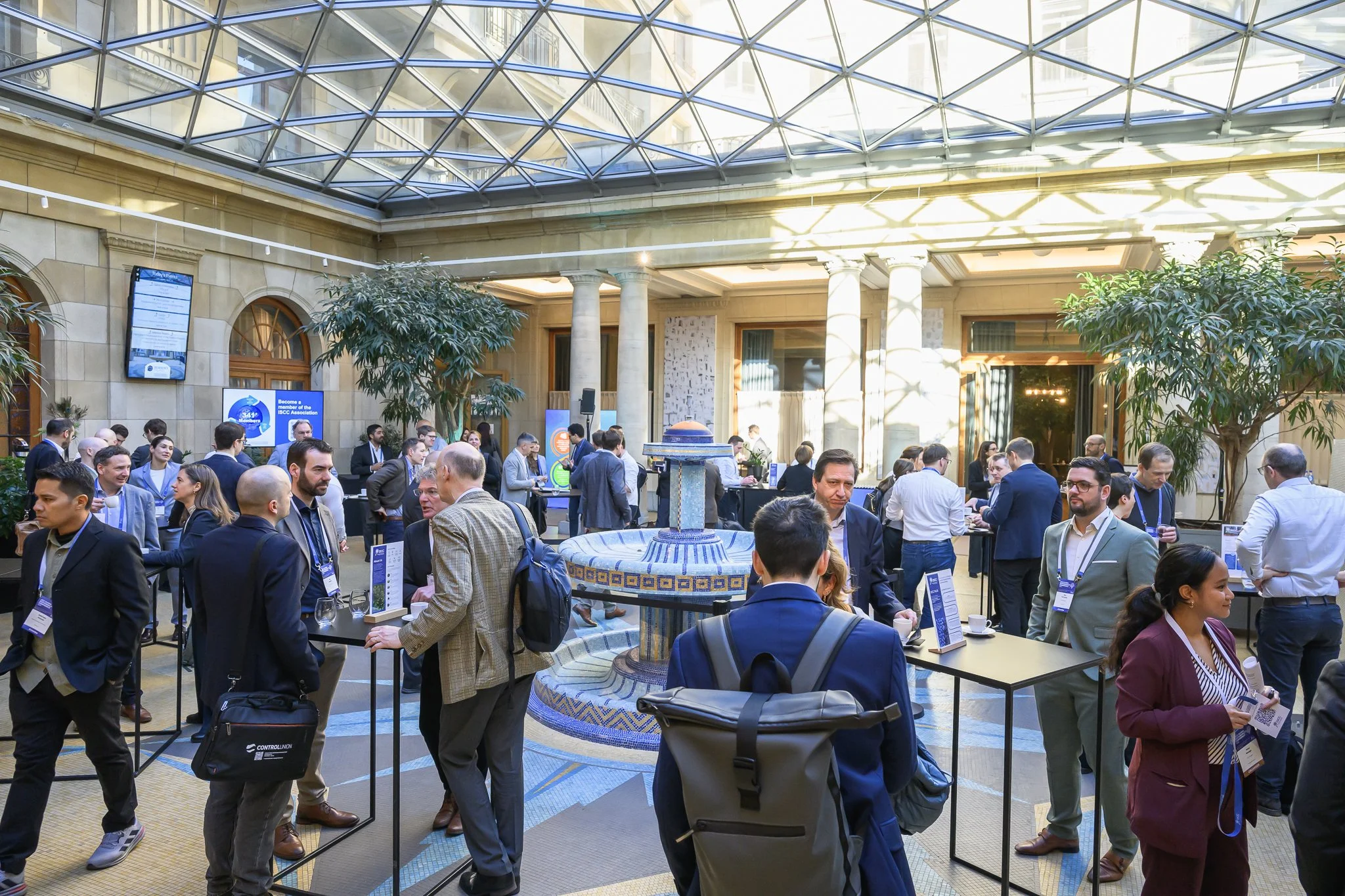 A wide view of participants networking around tall tables in a glass-roofed lobby.
