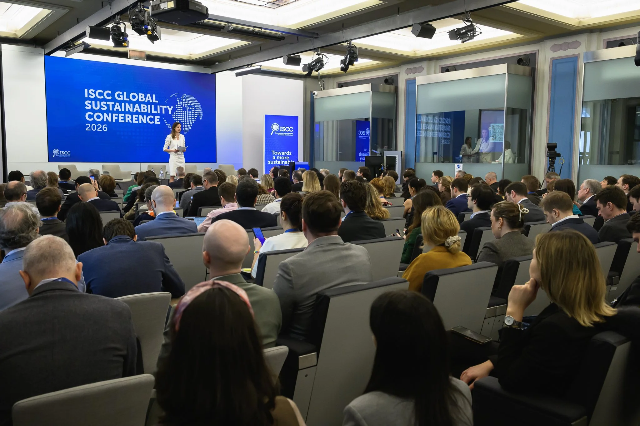 The conference audience faces the stage beneath ceiling-mounted lighting and projectors.