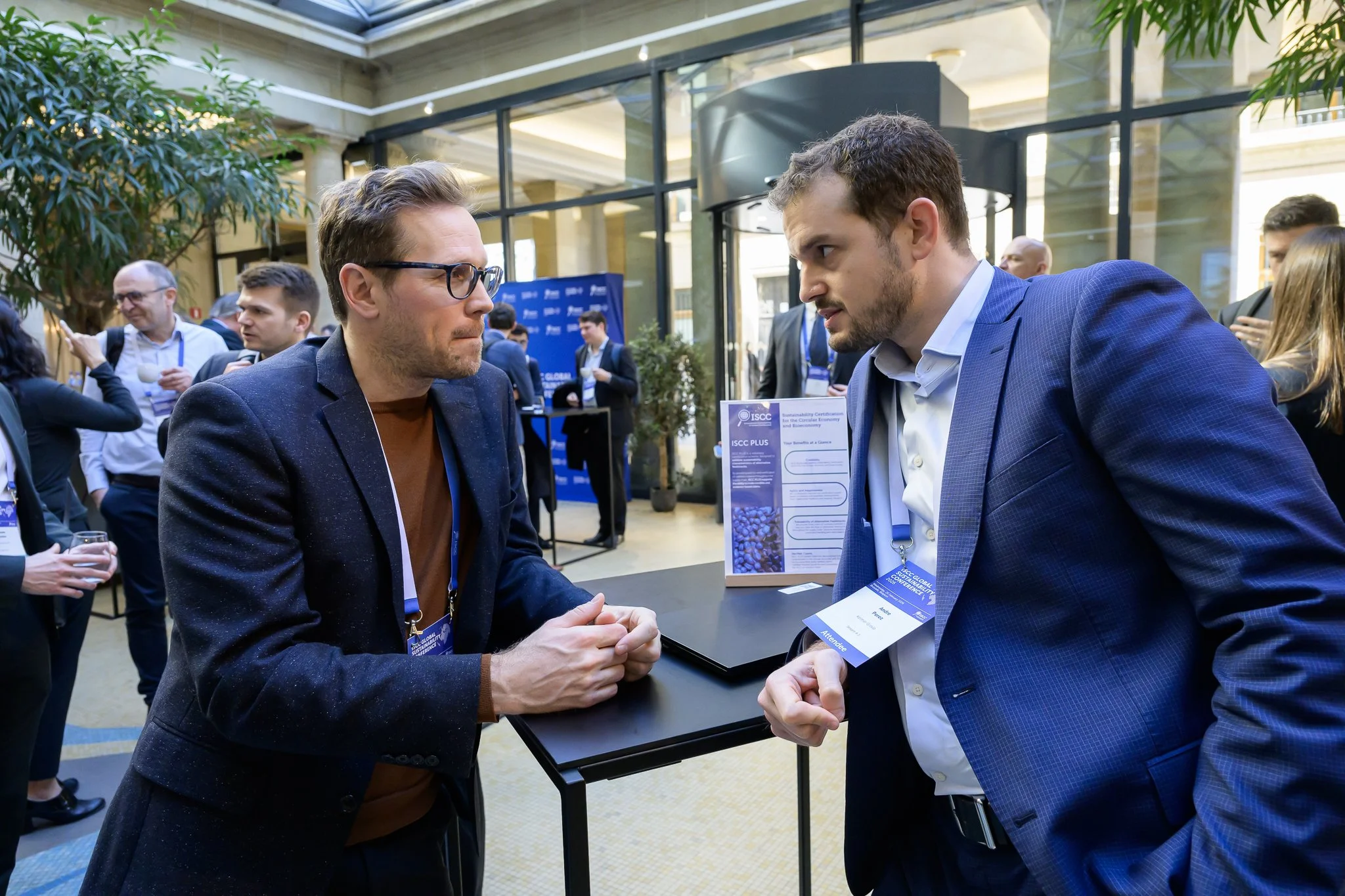 Two men lean towards each other while talking at a tall table.