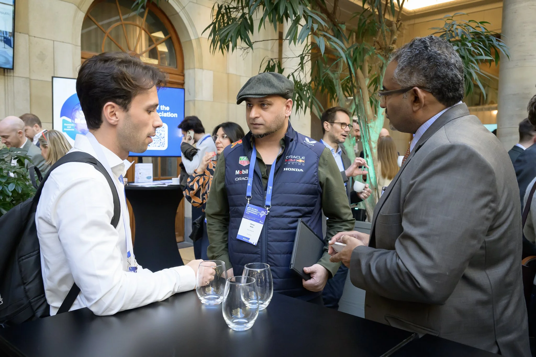 Three men talk around a tall table, with conference visitors behind them.